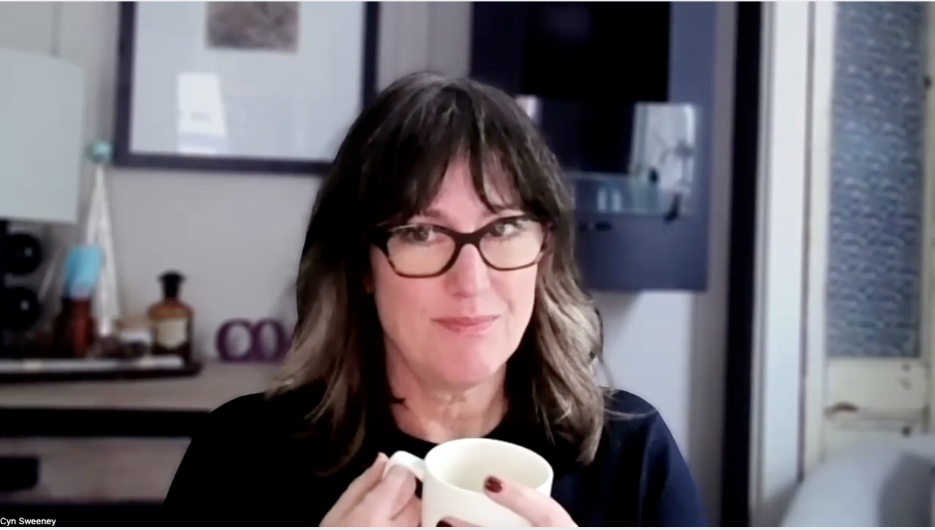 A woman with brown, mid-length hair and smiling, holds a coffee mug and glances to the side.